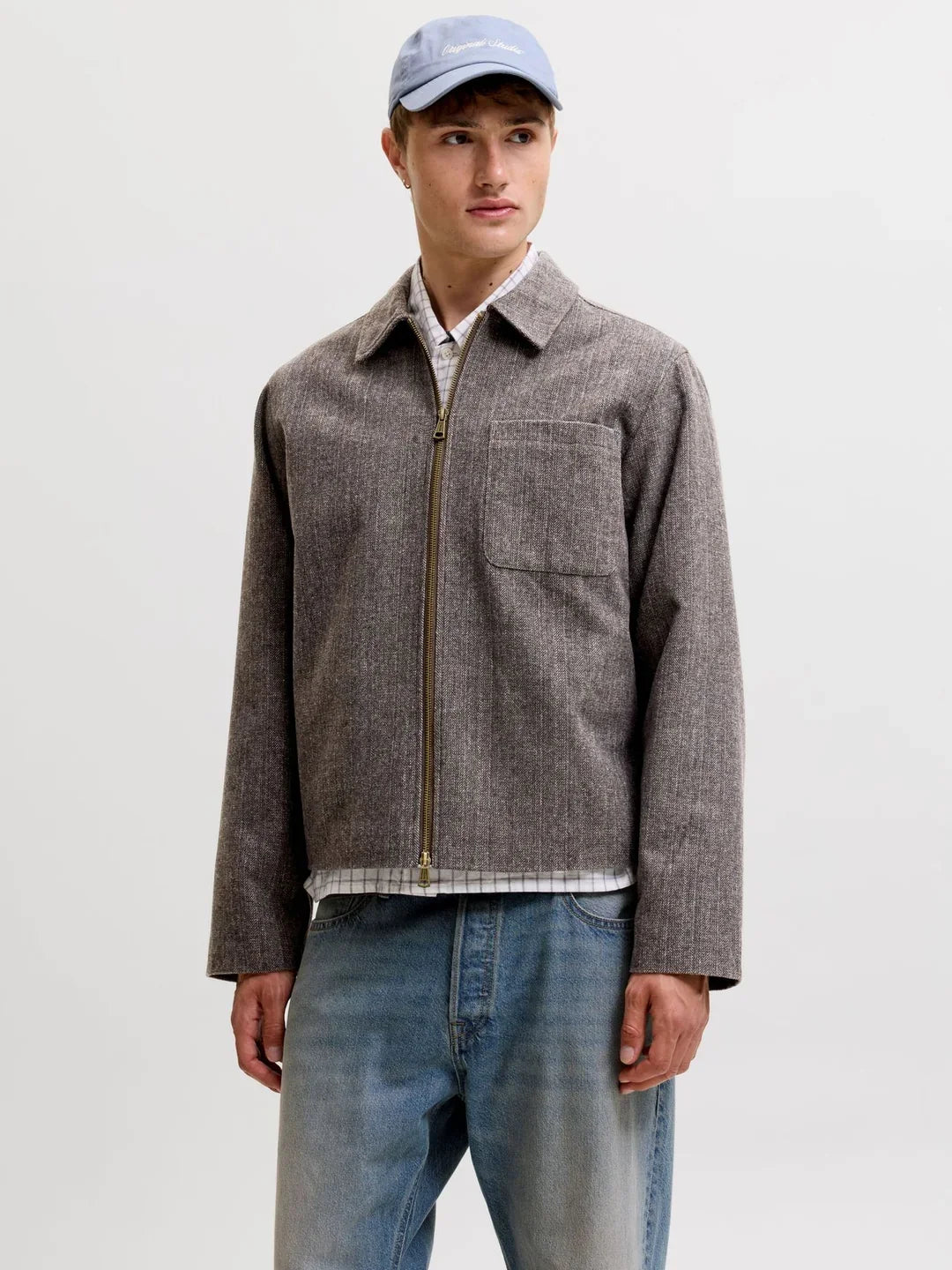 Model trägt eine braun-graue Herrenjacke mit Reißverschluss und Brusttasche über einem karierten Hemd. Eine helle Jeans und blaue Cap ergänzen den lässigen Look.