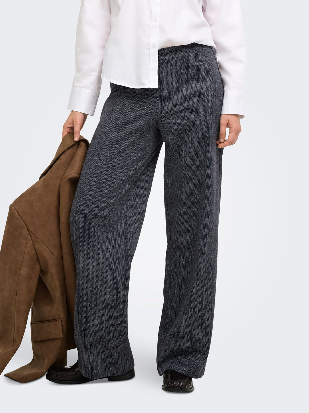 Model trägt eine dunkelgraue Wide-Leg-Hose mit feiner Fischgrätstruktur zu einer weißen Bluse. Ein brauner Blazer in der Hand ergänzt den eleganten Herbst-Look.