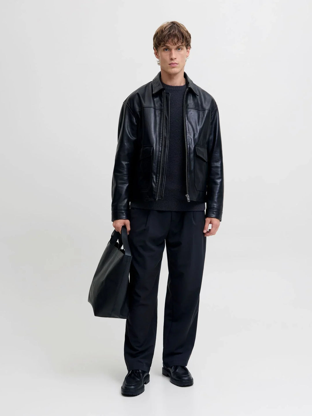 Model trägt eine schwarze Jacke in Lederoptik über einem dunklen Strickpullover und einer schwarzen Hose. Schwarze Schuhe und eine große Tasche ergänzen den modernen Ton-in-Ton-Look.