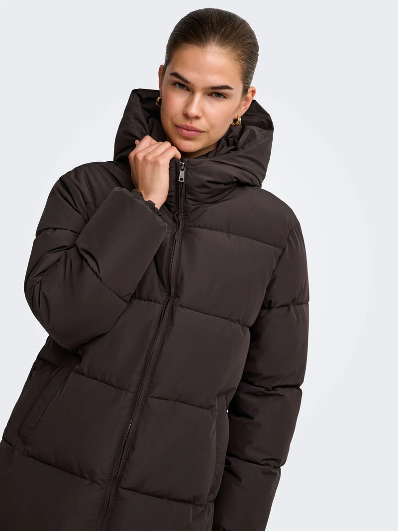 Model trägt einen dunkelbraunen, gesteppten Damenmantel mit großer Kapuze und durchgehendem Reißverschluss. Die Nahaufnahme zeigt den warmen, modernen Look für kühle Herbst- und Wintertage.